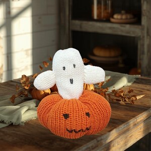 Pode incluir: Decoração de Halloween feita à mão com um fantasma branco em cima de uma abóbora laranja. O fantasma tem olhos e boca bordados em preto. A abóbora tem uma cara de lanterna esculpida. A decoração é feita de fio de malha e está numa mesa de madeira.