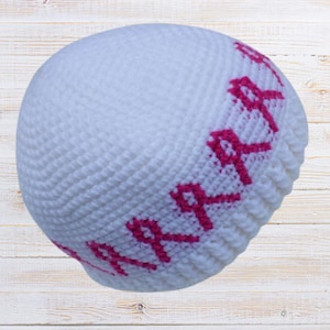 May include: A white crocheted beanie featuring pink ribbon designs around the lower edge. The hat is made of a soft, textured yarn and is designed to fit snugly. The pink ribbons are a prominent design element.