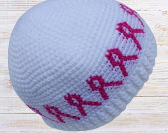 May include: A white crocheted beanie hat featuring a band of bright pink stitched lettering that reads "MARIA" in a repeating pattern around the lower section. The hat has a dense, textured knit construction with a ribbed edge at the bottom hem.