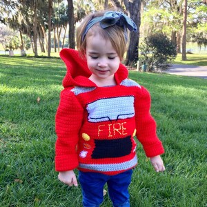 May include: A bright red crocheted hooded sweater featuring a fire truck design with a grey windshield, a black grille, yellow headlights, and the word FIRE embroidered in yellow thread across the front, paired with blue denim jeans.