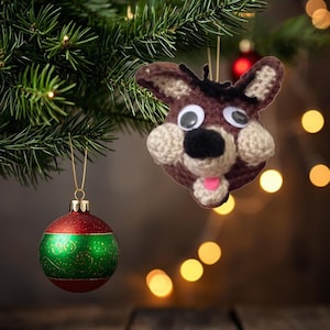 May include: A handmade Christmas ornament shaped like a dog's head, crafted from brown and beige yarn. The dog has googly eyes, a black nose, and a pink tongue. A red and green Christmas ball ornament hangs nearby.