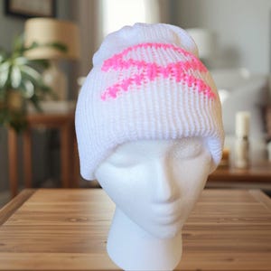 May include: White knitted beanie hat featuring a vibrant pink zigzag pattern across the top. The hat is displayed on a white mannequin head, highlighting the texture and design of the winter accessory.