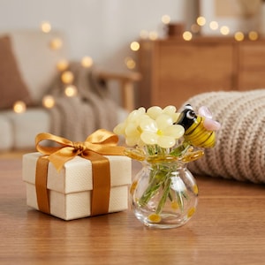 Puede incluir: Un pequeño jarrón de cristal transparente con flores de cristal amarillas y una figura de abeja rayada, junto a una caja de regalo cuadrada de papel crema texturizado, atada con un lazo ancho de cinta de satén dorado brillante.