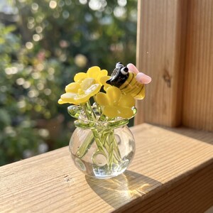 Puede incluir: Una escultura de vidrio en miniatura que presenta un jarrón redondo transparente con varias flores amarillas de tallos verdes y una pequeña abeja rayada con alas negras, amarillas y rosas posada sobre las flores, hecha de vidrio liso.