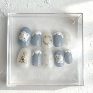 Pode incluir: Um conjunto de dez unhas artificiais numa caixa de apresentação transparente. As unhas apresentam desenhos com tema de inverno, incluindo bonecos de neve, árvores de Natal, estrelas douradas e uma grinalda dourada, com uma paleta de cores branca, azul claro e dourada.