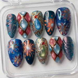 May include: A set of ten artificial nails with intricate designs. The nails feature a variety of colors, including blue, pink, and gold, with abstract patterns and embellishments. The nails are oval-shaped and displayed in a clear plastic case.