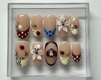 May include: A set of ten artificial nails in a clear plastic case. The nails feature various designs, including ladybugs, flowers, and polka dots. The color palette includes beige, red, white, and blue. Some nails have a French tip design.