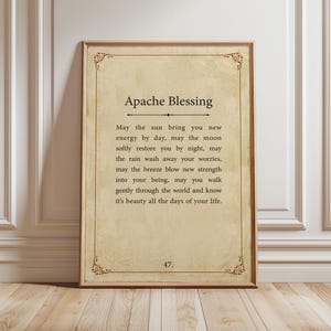 May include: A framed print with the text "Apache Blessing" in a decorative font. The text is on a beige background with a decorative border. The frame is a warm brown color. The print is displayed on a light wood floor.