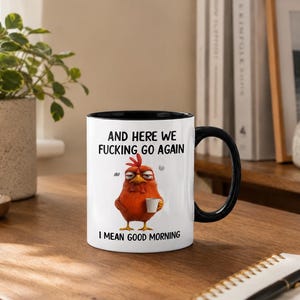 May include: White ceramic mug with a black interior and handle. The mug features the text "AND HERE WE FUCKING GO AGAIN I MEAN GOOD MORNING" and a cartoon chicken holding a coffee cup. The mug is on a wooden surface.