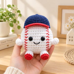 Può includere: Un peluche a forma di palla da baseball lavorato all'uncinetto in bianco con cuciture rosse, occhi in plastica nera e un piccolo sorriso ricamato. La palla indossa un cappellino da baseball blu scuro con visiera rossa e braccia.