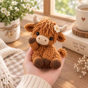 May include: A small, brown, crocheted highland cow plush toy with a cream-coloured muzzle, two textured horns, and a tiny white flower detail on its head. The cow has dark, round eyes and dark brown hooves, crafted with visible yarn stitches.