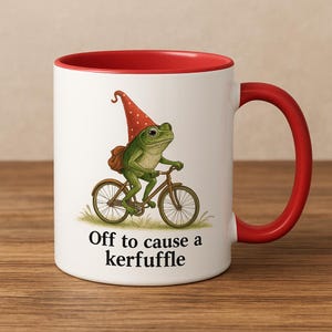 Könnte beinhalten: Weißer Keramikbecher mit rotem Innenraum und Henkel. Der Becher zeigt eine skurrile Illustration einer grünen Frosches auf einem Fahrrad, mit rotem Hut und Rucksack. Der Text "Off to cause a kerfuffle" steht darunter.
