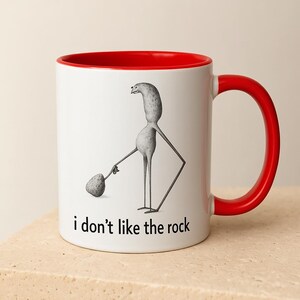 Puede incluir: Taza de cerámica blanca con interior y asa rojos. La taza presenta una ilustración caprichosa de una figura alta y delgada sosteniendo una roca, con el texto "i don't like the rock" impreso debajo. Adecuada para bebidas frías o calientes.