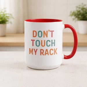May include: White ceramic mug with a red interior and handle. The mug features the text "DON'T TOUCH MY RACK" in colorful letters. The mug is a humorous kitchenware item.