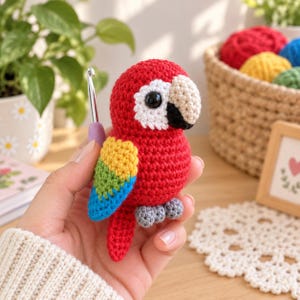 May include: A small, handmade crochet parrot toy featuring a vibrant red body, a white face patch, a beige beak with a black tip, and wings detailed with yellow, green, and blue yarn. The plush bird has small grey feet and a long red tail.