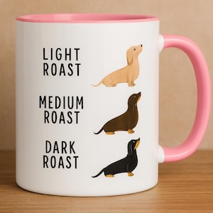 May include: White ceramic mug with a pink interior and handle. The mug features illustrations of dachshunds in light, medium, and dark colours, with the text "LIGHT ROAST", "MEDIUM ROAST", and "DARK ROAST" printed vertically on the mug.