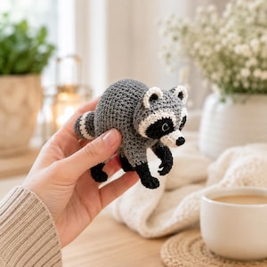 May include: A small, handcrafted crochet raccoon toy featuring a grey body, a striped tail, and a distinctive black and white facial mask. The figure has black paws, small rounded ears, and dark eyes, showcasing detailed yarn stitching throughout.