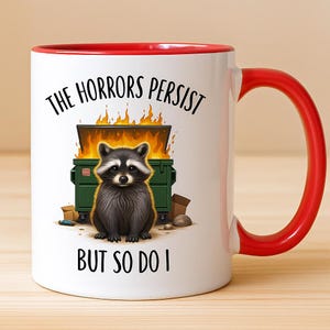 Peut inclure: Mug en céramique blanche avec intérieur et anse rouges. L'illustration représente un raton laveur devant un feu de poubelle, avec le texte "THE HORRORS PERSIST BUT SO DO I" en noir.