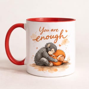 May include: White ceramic mug with a red interior and handle. The mug features a watercolor illustration of two hugging primates, with the text "You are enough" in a handwritten style. The design has a warm, comforting feel.
