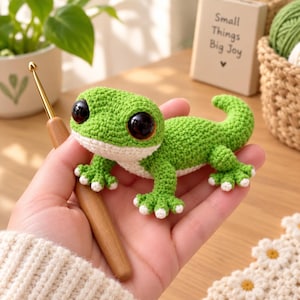 May include: A small, green, crocheted lizard toy with large, round, black eyes and a white underbelly. The lizard has four feet with white-tipped toes and a curved tail. A wooden-handled crochet hook is positioned next to the lizard.