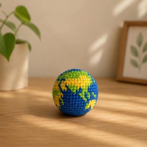 May include: A crocheted globe featuring blue oceans, green landmasses, and yellow continents. The handmade sphere sits on a wooden surface, showcasing the Earth's continents in a detailed, colorful design. The globe is a decorative item.