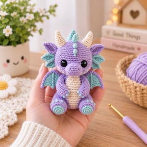 May include: A small, handcrafted crochet dragon toy featuring a purple body, cream-coloured belly, and light blue accents on the wings, horns, and feet. The creature has large black safety eyes, a rounded snout, and a series of small spikes along its back.