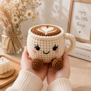 May include: A cream-colored crocheted mug with a brown heart-shaped latte art design on top. The mug features a smiling face with black eyes, eyelashes, and pink cheeks, along with two small brown feet at the base and a matching crocheted handle.