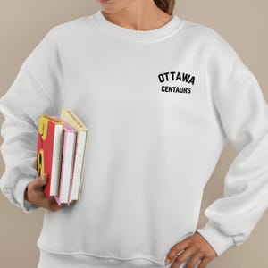 May include: White sweatshirt with the text "OTTAWA CENTAURS" in black. The person is holding three books with colorful covers. The sweatshirt has a crew neck and long sleeves.
