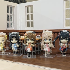 Op de afbeelding: Vijf anime-stijl beeldjes met grote ogen en kleurrijke outfits. De figuren staan op heldere plastic bases, geposeerd in een miniatuur kamer setting met een tafel en stoelen. De achtergrond heeft witte muren en houten accenten.