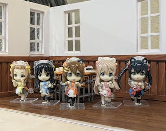 May include: Five anime-style figurines with large eyes and colorful outfits. The figures stand on clear plastic bases, posed in a miniature room setting with a table and chairs. The background features white walls and wooden accents.