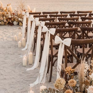 May include: Wedding ceremony setup on a sandy beach. Wooden chairs with white bows and ribbons line the aisle. Tall glass candle holders and floral arrangements with roses and pampas grass add to the decor. Soft, natural lighting.