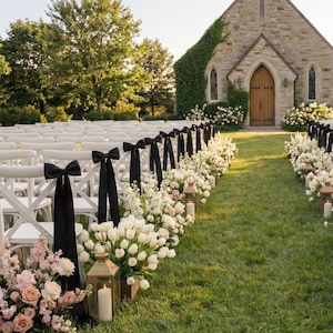 May include: Outdoor wedding ceremony setup with white chairs, black bows, and floral arrangements. The aisle is lined with white flowers, lanterns, and candles, leading to a stone building with a wooden door.