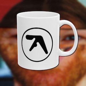 May include: White ceramic mug with a black logo inside a black circle. The logo is an abstract design. The mug has a curved handle. The background is blurred.