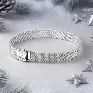 Könnte beinhalten: Ein silbernes Mesh-Armband mit rechteckigem Verschluss. Das Armband wird auf einer weißen Oberfläche mit silbernen Stern- und Tannenzapfendekorationen präsentiert. Der Verschluss trägt den eingravierten Markennamen.