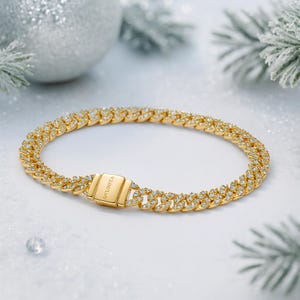 Puede incluir: Un brazalete dorado con diseño de eslabones cubanos, incrustado con pequeñas gemas transparentes. El brazalete tiene un cierre rectangular con la palabra "BALOOTA" grabada. La joya se exhibe sobre una superficie blanca y brillante.