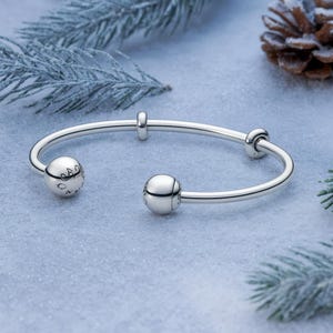 Può includere: Un bracciale a cerchio argentato con due terminali sferici e due piccoli elementi decorativi rotondi. Un terminale presenta dettagli incisi. Il bracciale è esposto su una superficie innevata con aghi di pino e una pigna sullo sfondo.