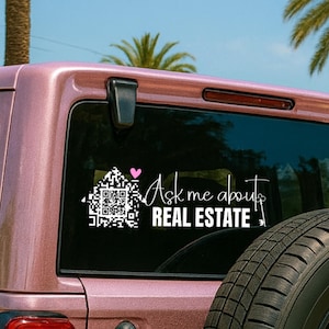 May include: A white vinyl decal on a vehicle's rear window. The decal features a QR code house graphic with a pink heart and the text "Ask me about REAL ESTATE" in a script font. The vehicle is a light pink color.