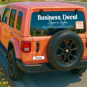 May include: An orange SUV with black wheels and a spare tire. The rear window displays a white decal that reads "Business Decal, Slogan or Tagline, Graphics | Logos | Custom" and a QR code. A small white sticker with the word "Custom" is on the bumper.