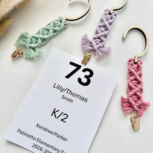 May include: Three macrame hangers in sage green, lavender, and dusty rose, each with a wooden hook and gold metal clip. A white rectangular card is attached, displaying the text 73, Lilly/Thomas Smith, K/2, Kendree/Parker, Palmetto Elementary School 2026-2027.