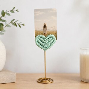 May include: A metal stand with a coiled base and a thin vertical rod topped with a light mint green macrame heart. The heart holds a rectangular photograph depicting a person in a hat standing in a field under a bright, cloudy sky.
