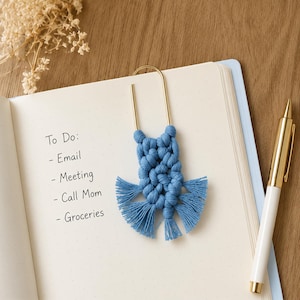 May include: A blue macrame bookmark attached to a gold-toned metal paperclip, resting on an open notebook with the text To Do: Email, Meeting, Call Mom, Groceries written on the page, accompanied by a white and gold pen.