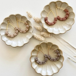 May include: Three pairs of handmade macrame earrings in shades of brown and tan, displayed on decorative scalloped plates. The earrings feature a teardrop shape with gold-colored metal hoops. Dried wheat stalks are arranged around the plates.