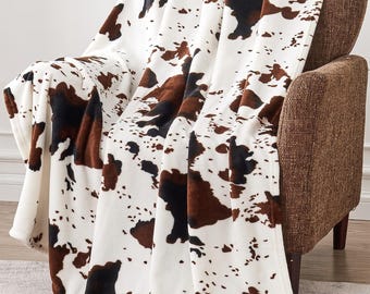 Op de afbeelding: Een zachte, witte deken met een koeienhuidpatroon in bruin- en zwarttinten is over een bruine fauteuil gedrapeerd. Het ontwerp van de deken heeft onregelmatige vlekken, wat een rustieke en gezellige uitstraling creëert. De deken is gemaakt van een pluche materiaal.