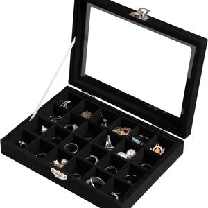 Puede incluir: Caja de joyería de terciopelo negro con tapa de cristal transparente, abierta para exhibir varios anillos y aretes. La caja está dividida en pequeños compartimentos para organizar joyas.
