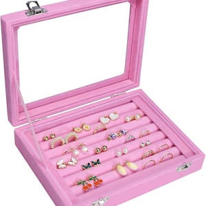 May include: A pink velvet jewelry box with a clear glass lid, open to display various earrings and rings. The box is filled with an assortment of earrings, including pearl studs, butterfly designs, and floral accents. Rings of various styles are also visible.
