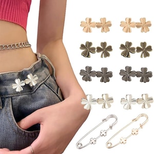 May include: A collection of clover-shaped brooches and safety pins in various metallic finishes. The brooches are in gold, silver, and gunmetal colors. The safety pins have clover accents. The brooches are shown attached to denim.