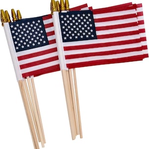 May include: A set of small handheld United States flags, each featuring a blue canton with white stars and alternating red and white horizontal stripes. The flags are attached to light-colored wooden poles topped with small, pointed gold-colored finials.