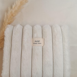 Pode incluir: Uma clutch retangular branca de pele sintética com detalhes canelados verticais. Uma pequena etiqueta retangular no centro diz "AVAÉ Family Couture". A clutch é um acessório macio e fofo.