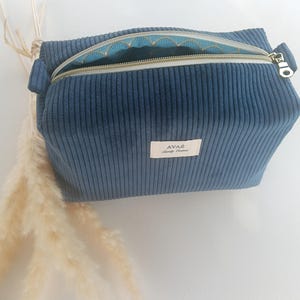 Peut inclure: Une trousse de maquillage en velours côtelé bleu avec une fermeture éclair dorée et un intérieur à motifs. Le sac est rectangulaire et porte une étiquette "AVAE". La fermeture éclair est ouverte.