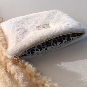 Könnte beinhalten: Eine flauschige, weiße Clutch mit goldenem Reißverschluss und Leopardenmuster-Futter. Die Clutch hat ein kleines rechteckiges Etikett mit Text. Hergestellt aus weichem, plüschigem Material.
